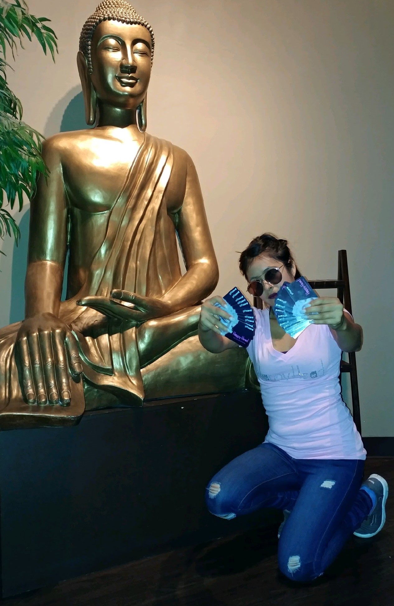Person posing in front of a large golden Buddha statue at Happy Head Massage, holding multiple gift cards, symbolizing generosity and relaxation offerings.
